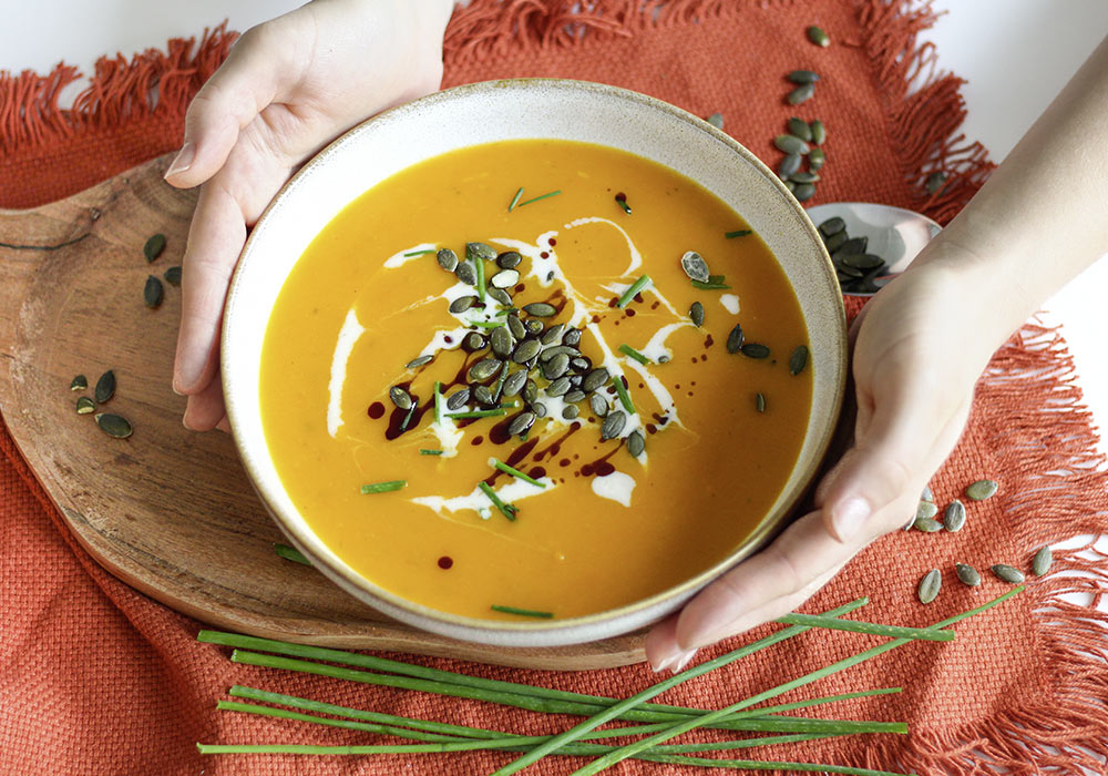 Gelbe Kürbissuppe mit verschiedenen Toppings in einer grauen Porzellanschüssel. Darunter ein Holzbrett und ein rotes Leinentuch. Außerdem sieht man Kürbiskerne und Schnittlauch verteilt. 