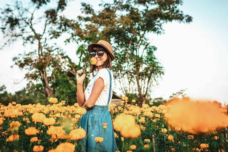 Frau mit Hut steht in einem Blumenfeld und riecht an einer Blume