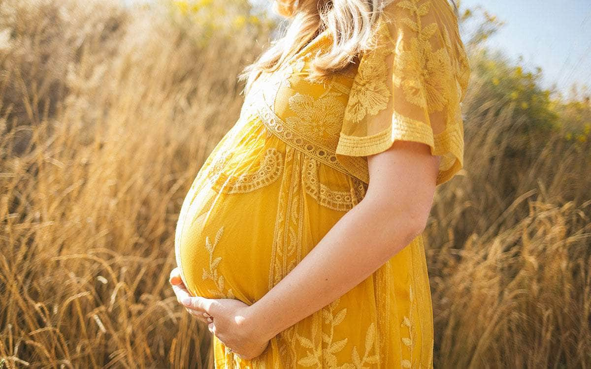 Eine schwangere Frau im gelben Kleid steht vor einem Getreidefeld und umfasst ihren Bauch.