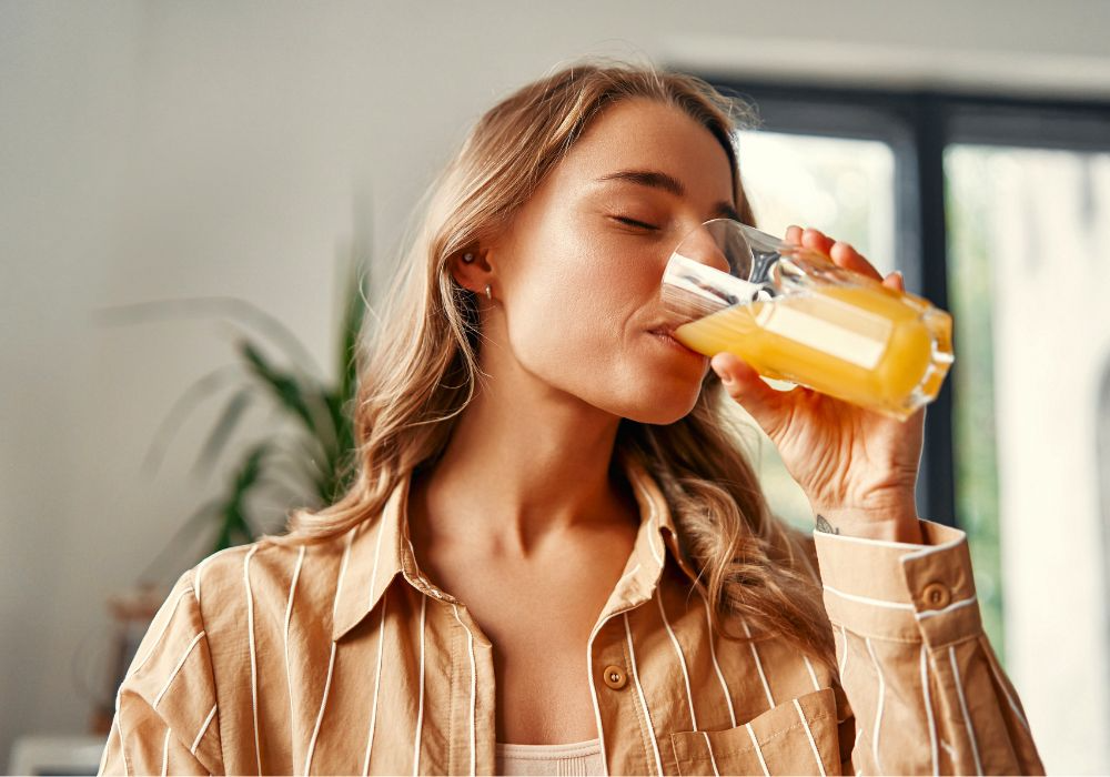 Person mit langen mittelblonden Haaren und braunem Hemd trinkt Glas Saft