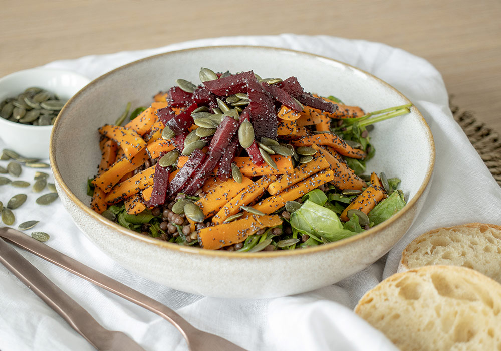 Linsen-Ofengemüse-Salat mit Mohn und Pflaumendressing und Ciabatta Brot auf einer weißen Tischdecke. Im Hintergrund sind Kürbiskerne zu sehen.
