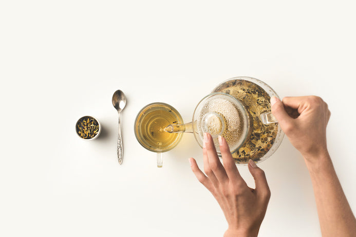 teeloeffel-teetasse-und-loser-tee-liegen-auf-einem-weißen-tisch-frauenhaende-halten-teekanne-aus-glas-und-gießen-tee-in-tasse
