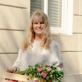 Julia von InnoNature, eine blonde Frau, steht mit einem Korb voller frischem Gemüse vor einer Hauswand und lächelt in die Kamera.