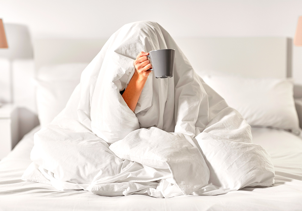 Person sitzt auf einem Bett mit der Decke über dem Kopf und einer Tasse in der Hand.