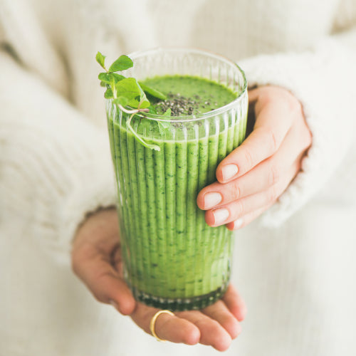 Grüner Smoothie wird von Frau mit beigem Pullover gehalten