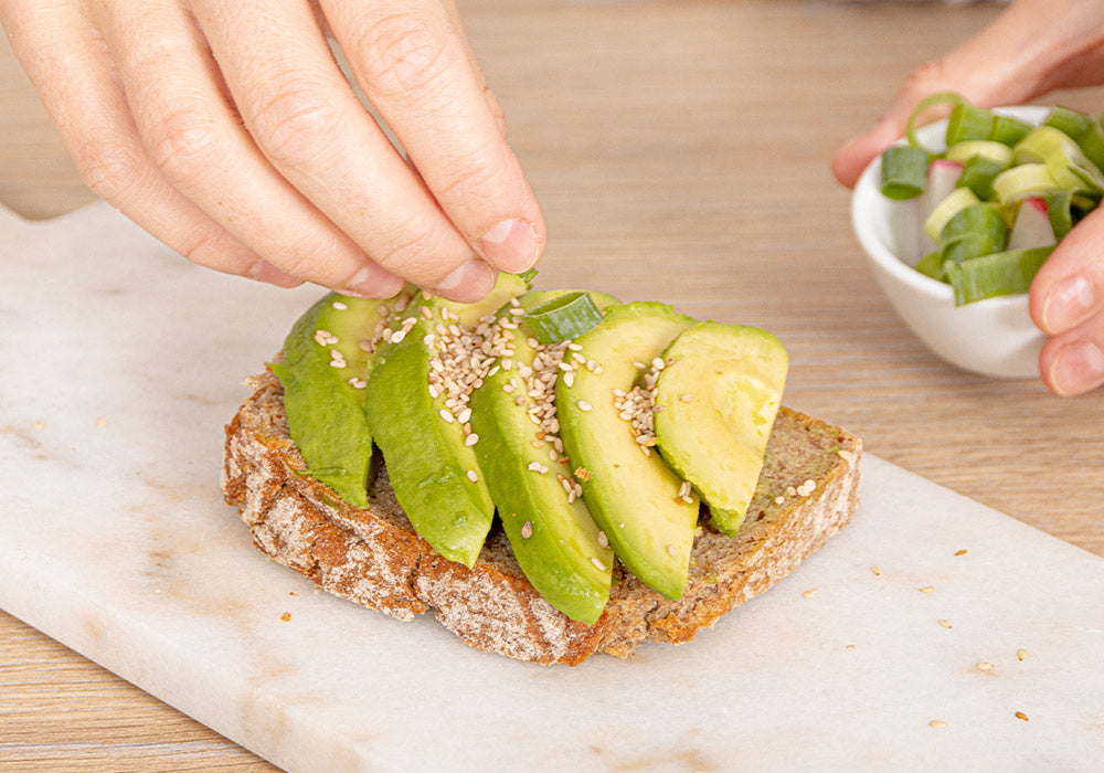 Hände träufeln Sesam auf ein Avocadobrot.