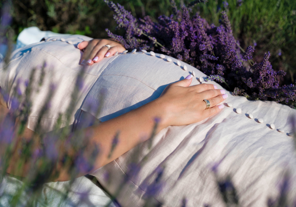 Eine schwangere Frau im gelben Kleid steht vor einem Getreidefeld und umfasst ihren Bauch.