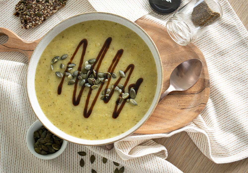 Schnelle Fenchel Suppe dekoriert mit Kürbiskernen und Balsamico auf einem Holzbrett. Darunter liegt eine weiß-braune Tischdecke. Drumherum sind Gewürze und ein Stück Brot zu sehen.