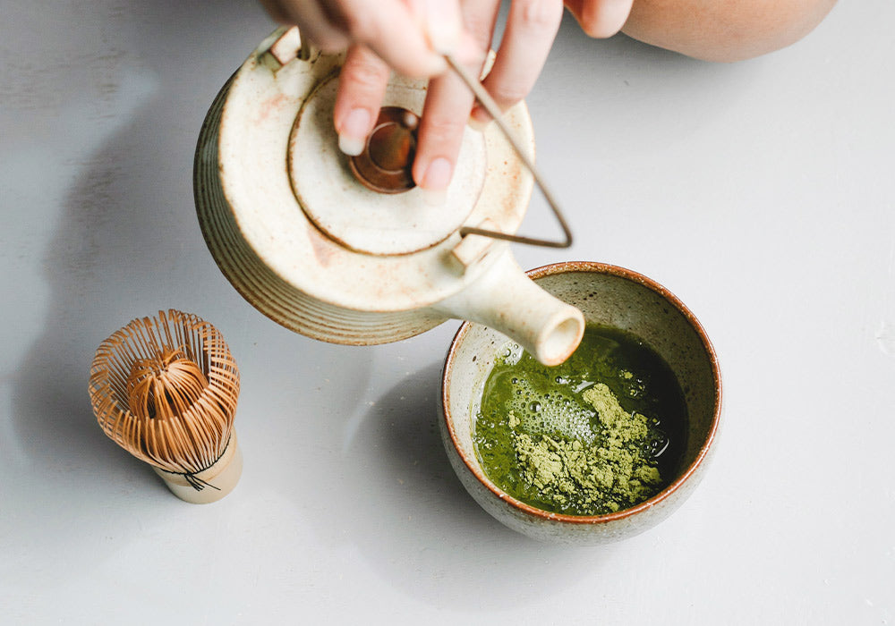 Eine Frauenhand hält eine Teekanne und gießt Matcha Tee auf.