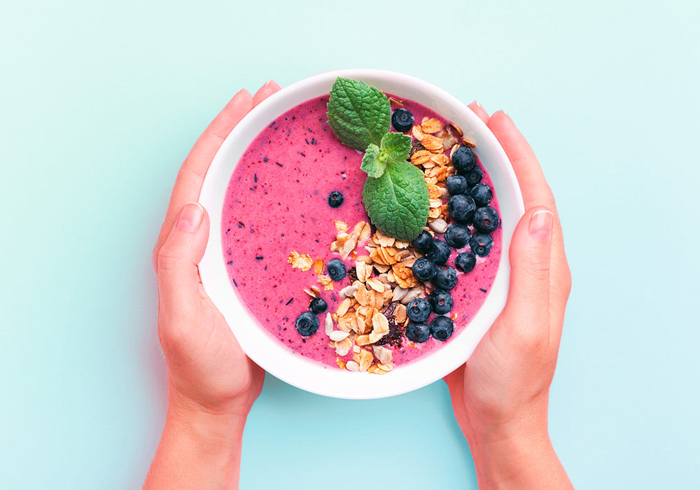Hände halten eine Smoothiebowl mit Blaubeeren als Topping vor hellblauem Hintergrund