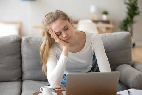 Frau sitzt auf grauer Couch vor einem Laptop und hält sich den Kopf, als wäre sie müde