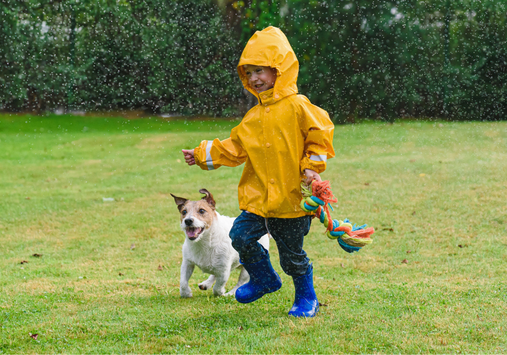 Kind springt mit einem Hund bei Regen vergnügt über eine Wiese