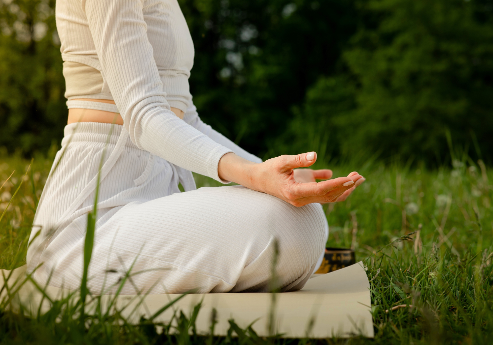 Frau mit rosafarbenem Oberteil sitzt im Schneidersitz, einer Meditationspose und hält ihre Hände zu einem Mudra – Daumen und Zeigefinger berühren sich jeweils – auf ihren Knien.