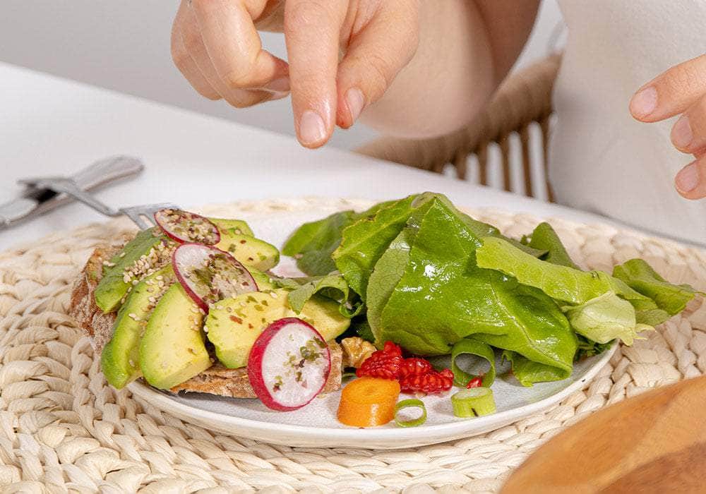 Auf einem Teller ist ein grüner Salat und ein Avoadobrot mit Radieschen, Möhre, Himbere und Sesam garniert.