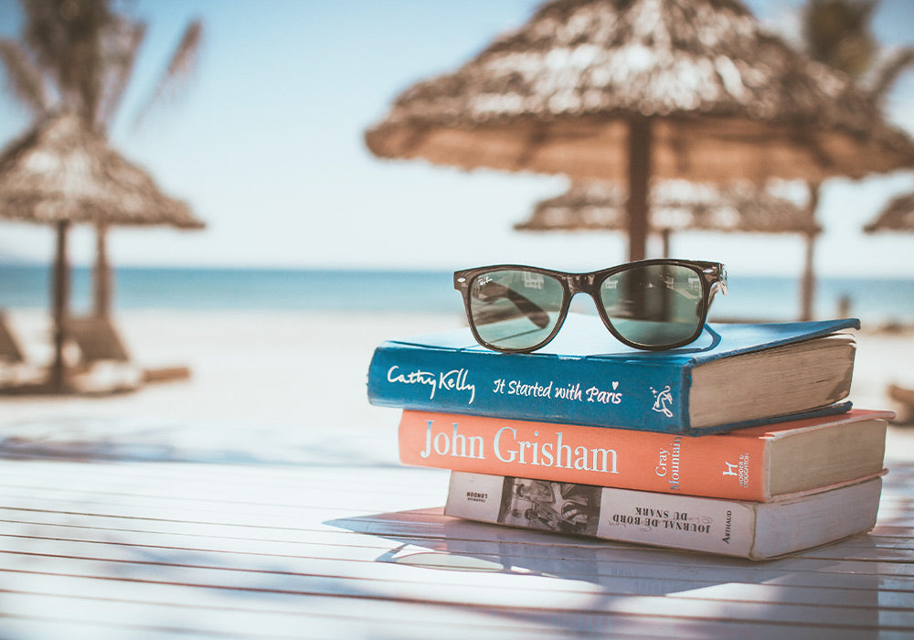 Am Strand liegt ein Stapel mit Büchern, oben drauf eine Sonnenbrille.