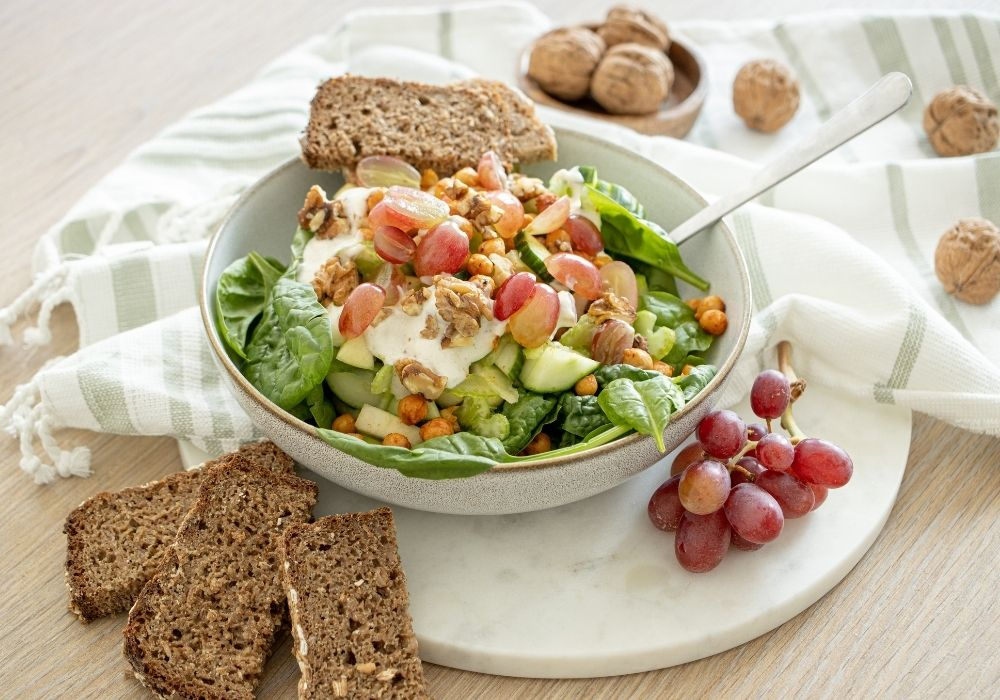 Leichter Waldorf-Salat mit gerösteten Kichererbsen in einer grauen Porzellan Schüssel. Drumherum sind Weintrauben, Walnüsse und drei kleine Scheiben Vollbrot zusehen. 
