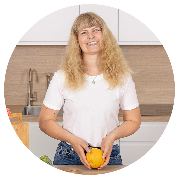 Bild einer jungen Frau mit blonden Haaren und weißem T-Shirt. In der Hand hält sie eine Orange. 