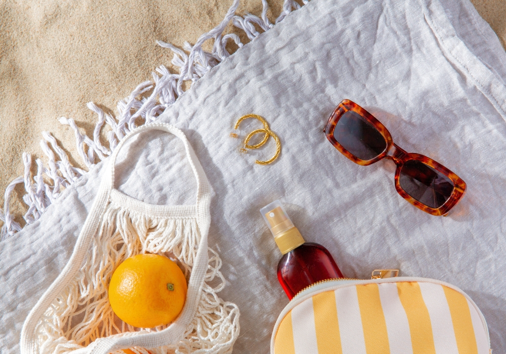 Weißes Strandtuch auf Sand mit Häkeltasche, Sonnenbrille, Sonnenöl, Orange und Ohrringen