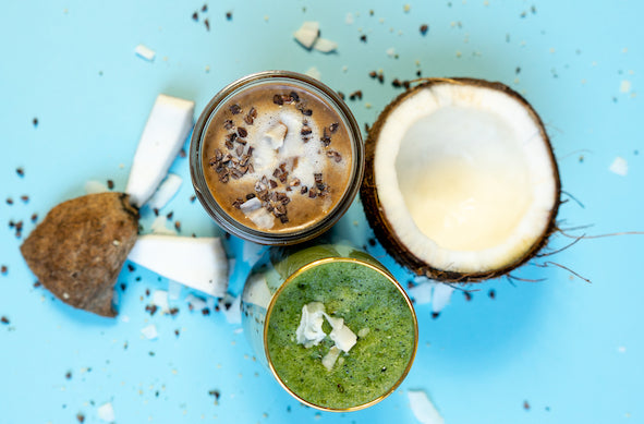 zwei-glaeser-eins-mit-braunem-shake-eins-mit-gruenem-shake-daneben-kokosnusshälften-und-kakaonibs-auf-blauem-untergrund