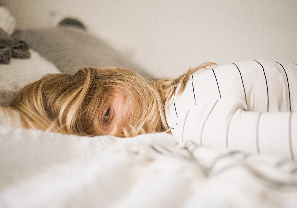Blonde Frau liegt auf dem Bett, die Haare verdecken das Gesicht und sie sieht müde aus. 